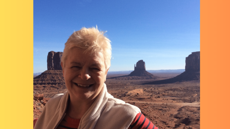 the author Charley Linden Thorp stands in the foreground smiling against a backdrop of Monument valley graced with a stunning clear blue sky